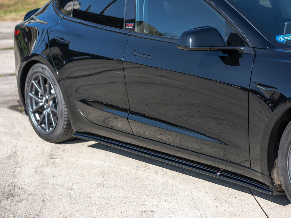 A black car is parked on a concrete surface. The side section, including the door and rear wheel, is visible. A sticker on the window reads "TESLA."