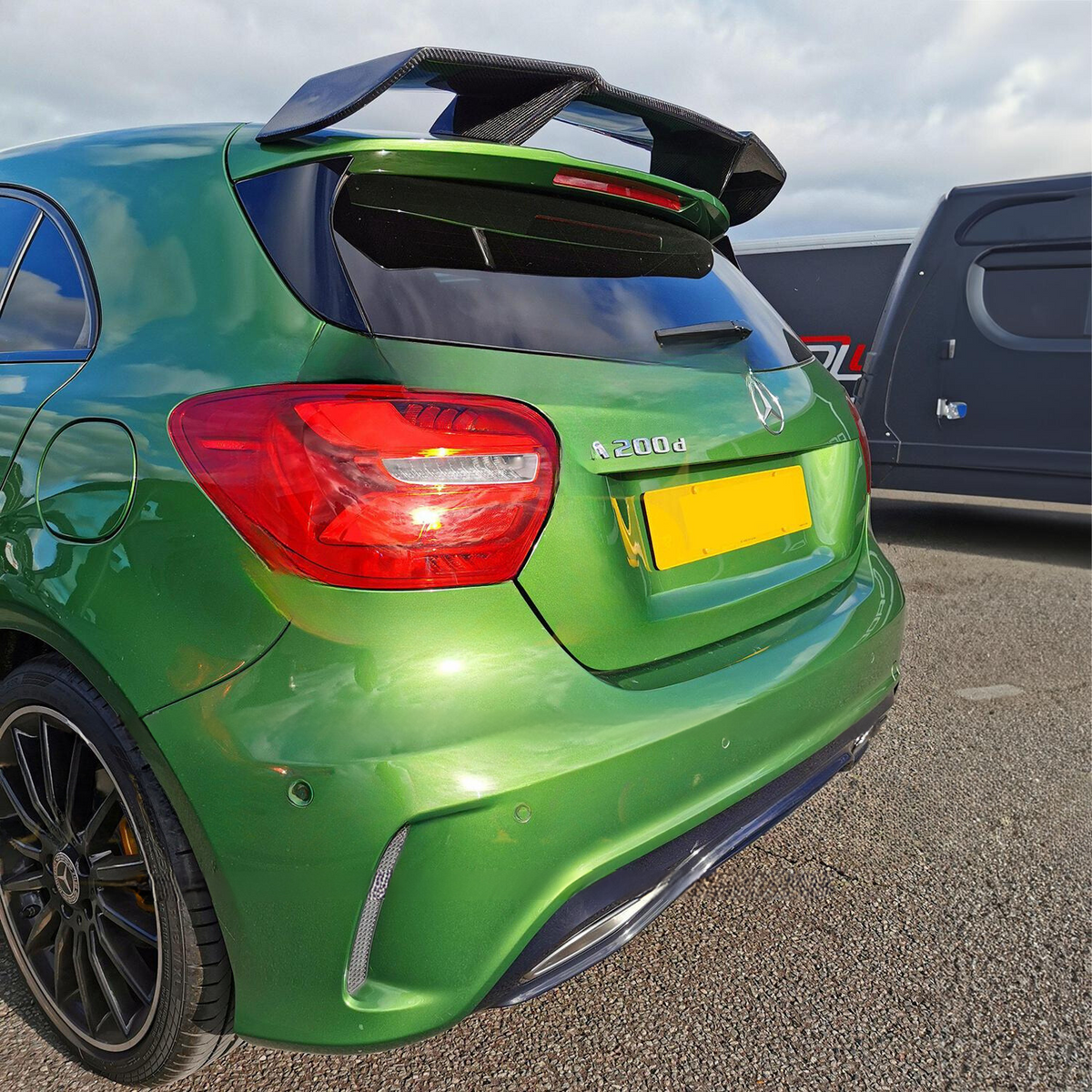 Green Mercedes A 200 d car parked with a spoiler and tinted windows visible, surrounded by a gray vehicle on an asphalt surface. License plate is obscured.