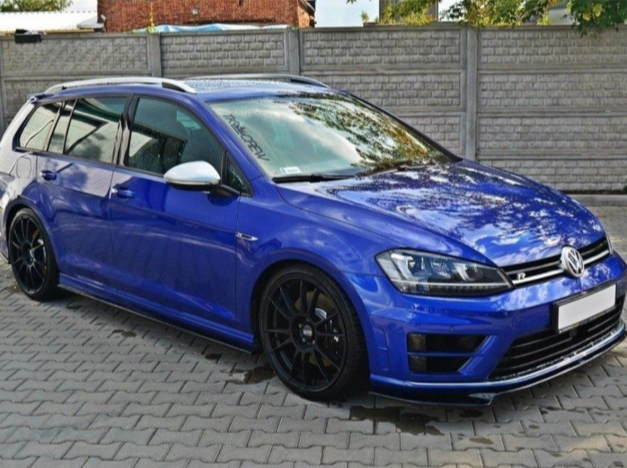 A blue Volkswagen station wagon is parked on a stone-paved area, surrounded by a brick wall and trees. The windshield displays text: "RACINGTEAM."