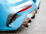 The rear section of a blue sports car with carbon fiber detailing features dual exhaust pipes, a red reflector, and is parked on a paved surface.