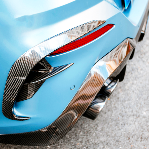 The rear section of a blue sports car with carbon fiber detailing features dual exhaust pipes, a red reflector, and is parked on a paved surface.