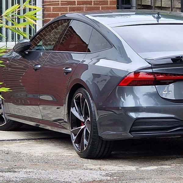 A sleek, gray car is parked on a driveway next to a brick building and wooden fence, with plants on the left side.