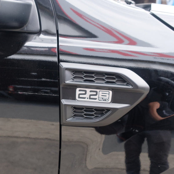 A silver vehicle emblem reads "2.2 6 AUTO" with a honeycomb design, mounted on a glossy black car surface, reflecting faint surroundings in an outdoor setting.