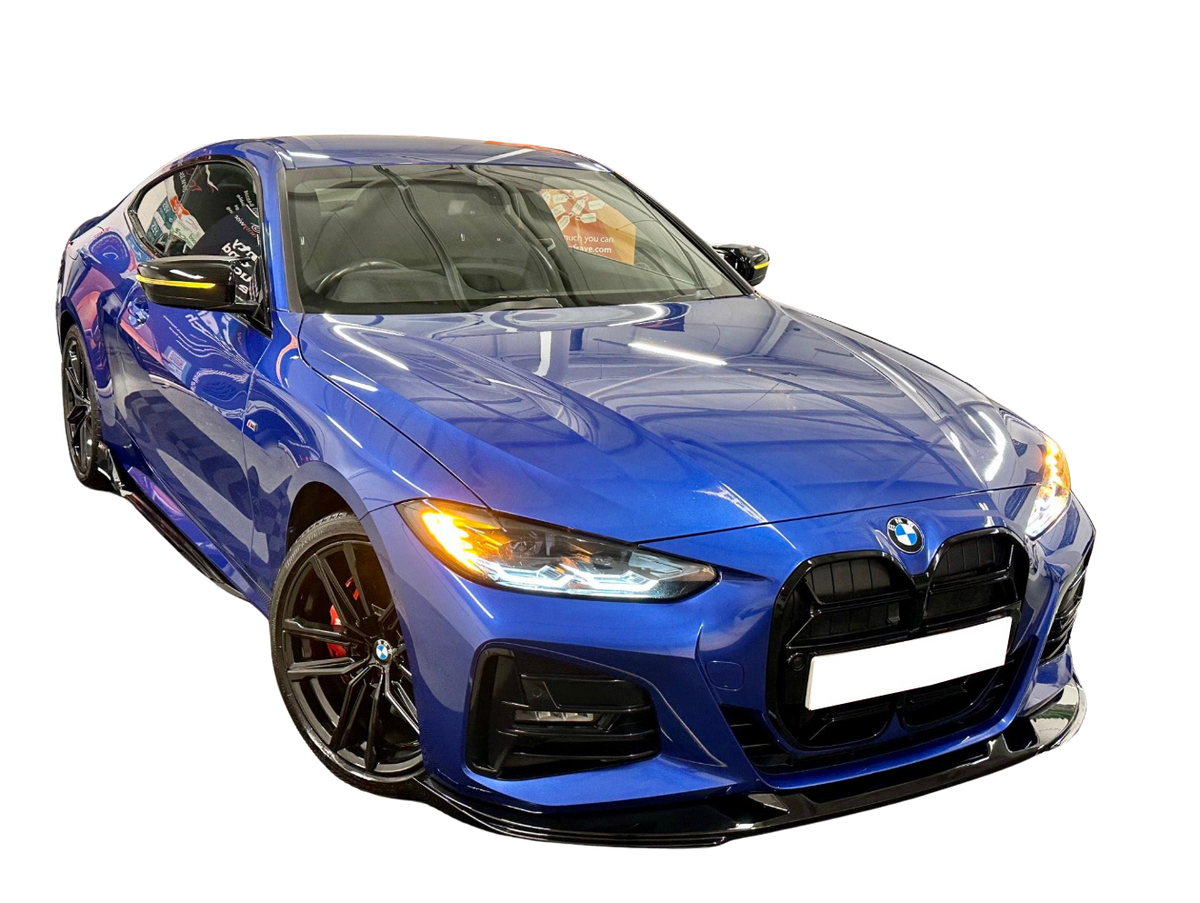 A blue sports car with headlights on is parked in a showroom, reflecting overhead lights. The environment's reflections highlight the vehicle's sleek, aerodynamic design.