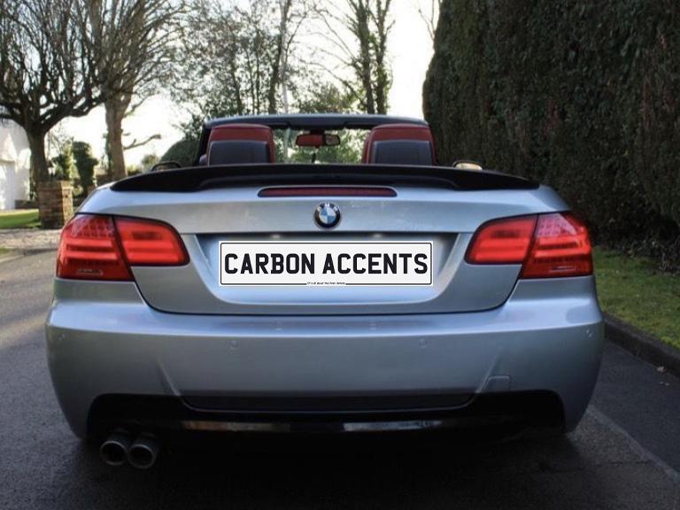 A silver convertible car, parked on a road, displays the license plate text "CARBON ACCENTS." Trees and hedges line the background, suggesting a suburban environment.