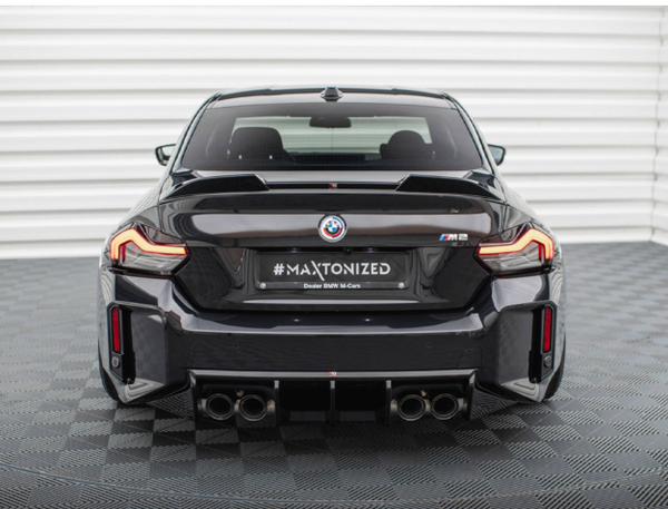 A black BMW M2 displayed in a showroom with checkered flooring. The car's rear features quad exhausts and illuminated taillights. Text on the license plate reads "#MAXTONIZED Dealer BMW M Cars."
