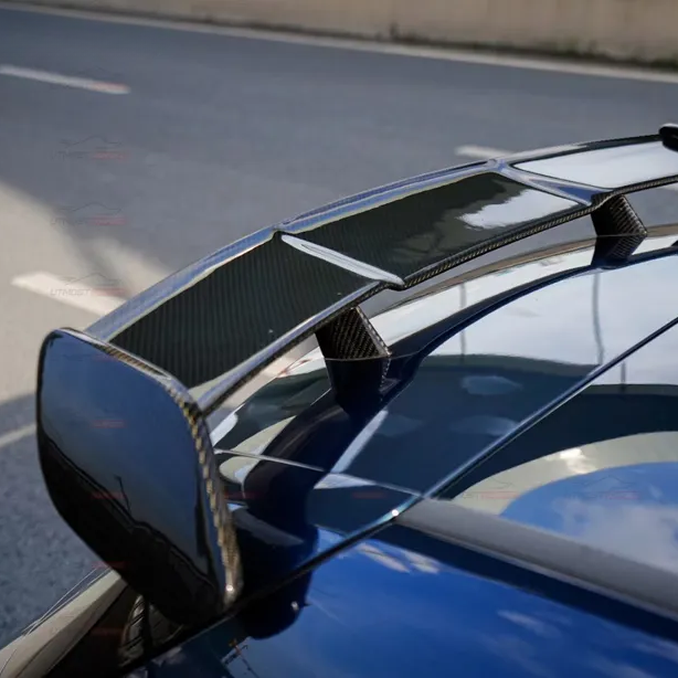 A carbon fiber car spoiler sits on a blue vehicle, reflecting the sky. It's positioned beside a road with a concrete barrier, suggesting a street-setting.
