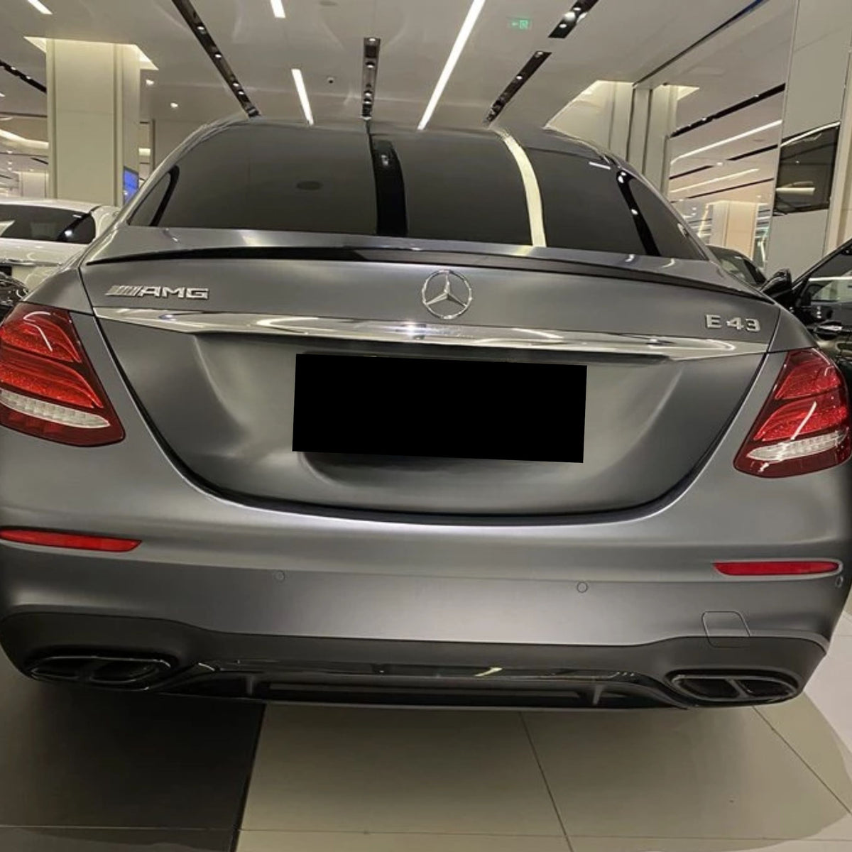 A gray Mercedes-Benz E43 AMG sedan is parked indoors, with its rear facing the viewer. Bright showroom lights reflect on the car's body.