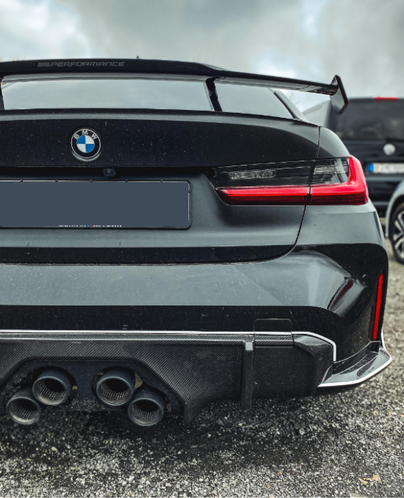 The rear view of a black BMW car with a visible logo, quad exhausts, a "M Performance" spoiler, and red taillights, is parked on gravel with cloudy skies overhead.