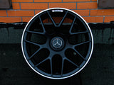 A black Mercedes AMG car wheel, featuring a glossy emblem at the center, rests against an orange brick wall on a textured gray surface.