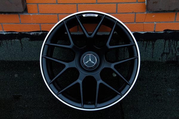 A black Mercedes AMG car wheel, featuring a glossy emblem at the center, rests against an orange brick wall on a textured gray surface.