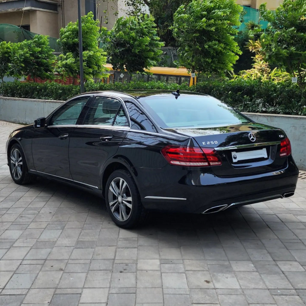 A black Mercedes E250 is parked on a tiled driveway, surrounded by lush green bushes and urban buildings, reflecting the sunlight on its polished surface.