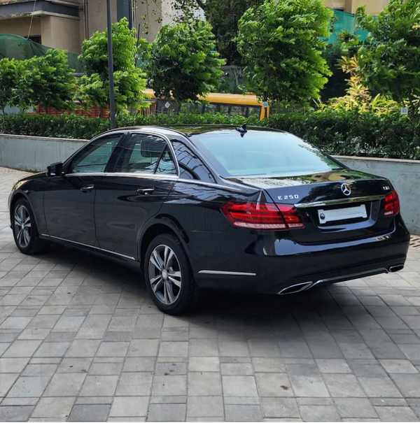 A black Mercedes E250 is parked on a tiled driveway, surrounded by lush green bushes and urban buildings, reflecting the sunlight on its polished surface.