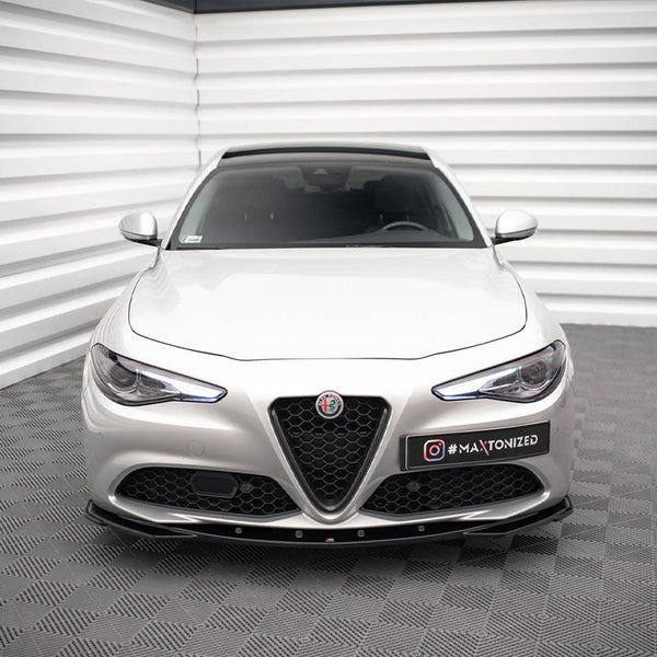 A silver car is parked in a modern showroom with textured walls and flooring. The car's front features a distinctive emblem and a license plate reading "#MAXTONIZED."