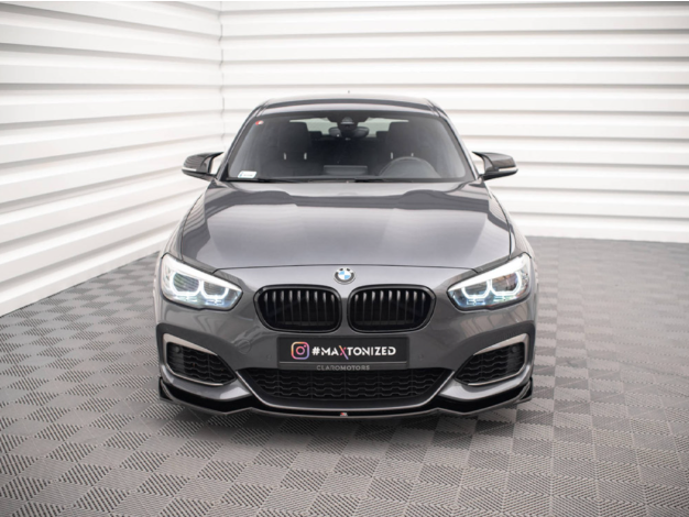 A gray BMW car is parked on a patterned surface, facing forward. Text on the front reads "#MAXTONIZED CLEATONMOTORS." The background shows a white, ridged wall.
