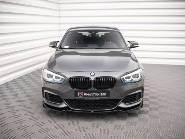 A gray BMW car is parked on a patterned surface, facing forward. Text on the front reads "#MAXTONIZED CLEATONMOTORS." The background shows a white, ridged wall.