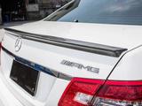 A white car with AMG and Mercedes logos features a carbon fiber spoiler on the trunk lid, parked near a building with various objects in the background.