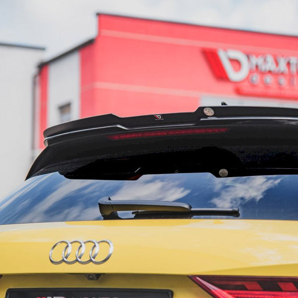Yellow Audi parked with rear view featuring a spoiler and license plate. Behind, a red building displays the text "MAXTON design." The sky is partly cloudy.