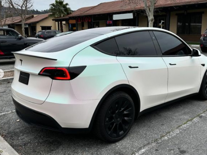 A white car with tinted windows and black wheels is parked in a lot. Surroundings include other vehicles and buildings with signs such as "TATTOO" visible.