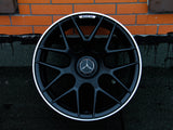 A black alloy wheel with a Mercedes emblem rests against an orange brick wall. The wheel features a white rim trim and 