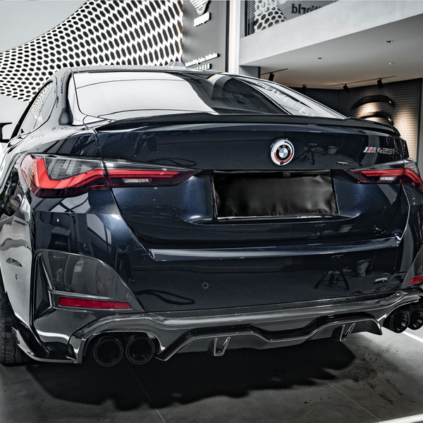 A sleek, dark-colored car is parked in a showroom with a modern design. The car's rear features prominent taillights and quad exhaust pipes. The wall has text: "wrkz DryCarbon™."