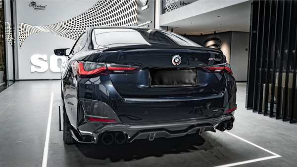 A sleek, dark-colored car is parked in a showroom with a modern design. The car's rear features prominent taillights and quad exhaust pipes. The wall has text: "wrkz DryCarbon™."