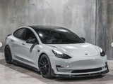 A white Tesla car is parked in a concrete-walled room, displaying a sleek, modern design with black-rimmed wheels and tinted windows.
