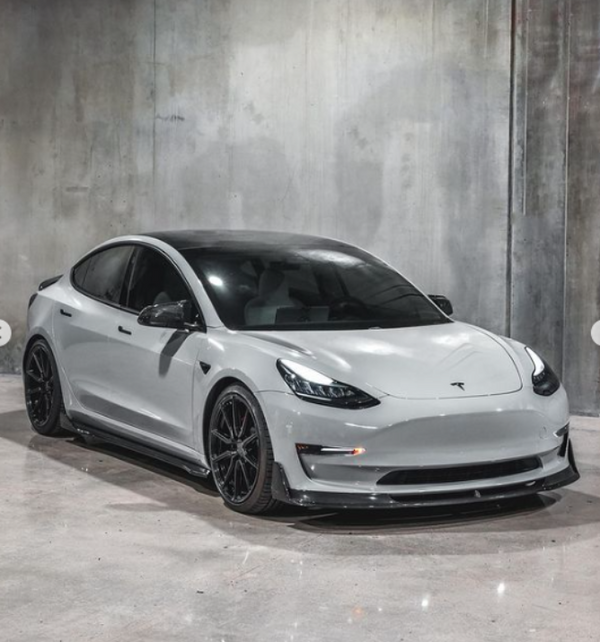 A white Tesla car is parked in a concrete-walled room, displaying a sleek, modern design with black-rimmed wheels and tinted windows.
