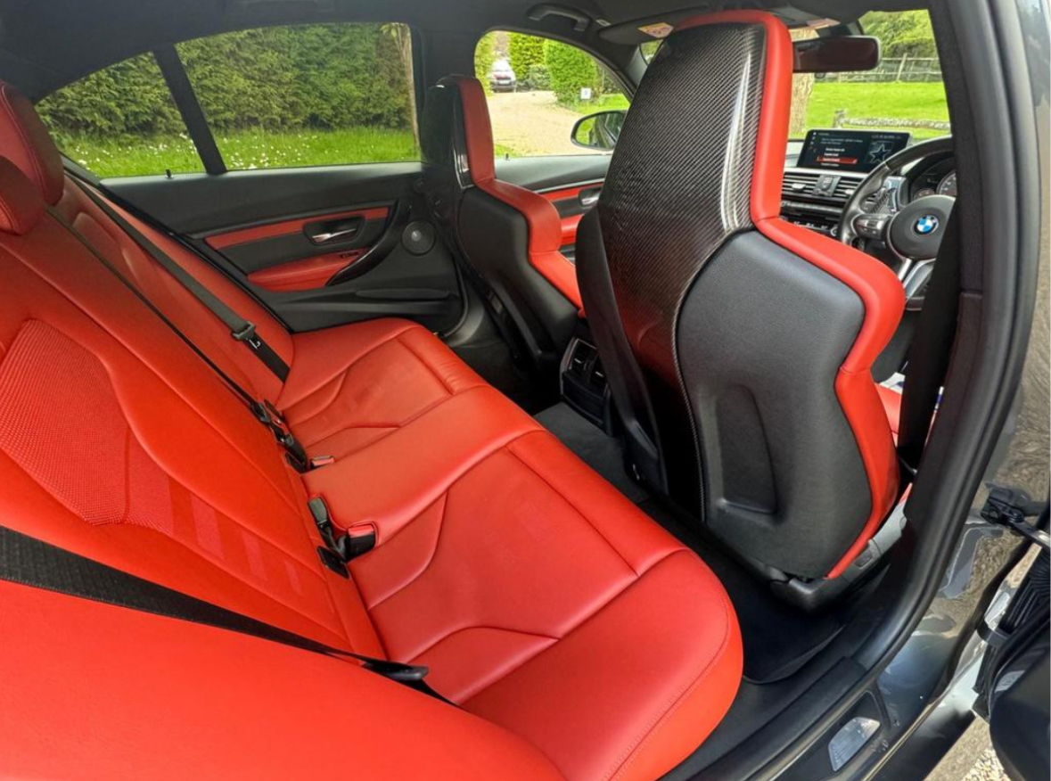 Red leather seats are positioned in the rear of a car with a sleek, modern interior. The door is open, revealing a grassy outdoor area.