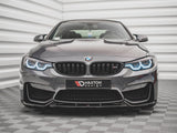 A sleek, dark BMW with illuminated headlights sits on a patterned floor, facing forward. Behind it is a textured, white wall. The front license plate reads 