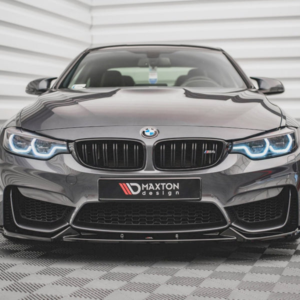 A sleek, dark BMW with illuminated headlights sits on a patterned floor, facing forward. Behind it is a textured, white wall. The front license plate reads "MAXTON design."