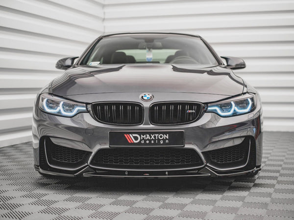 A sleek, dark BMW with illuminated headlights sits on a patterned floor, facing forward. Behind it is a textured, white wall. The front license plate reads "MAXTON design."