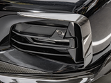 Close-up of a car’s front bumper featuring a sleek, black grille with carbon fiber trim detailing; the reflection and lighting highlight the material's texture in an indoor setting.