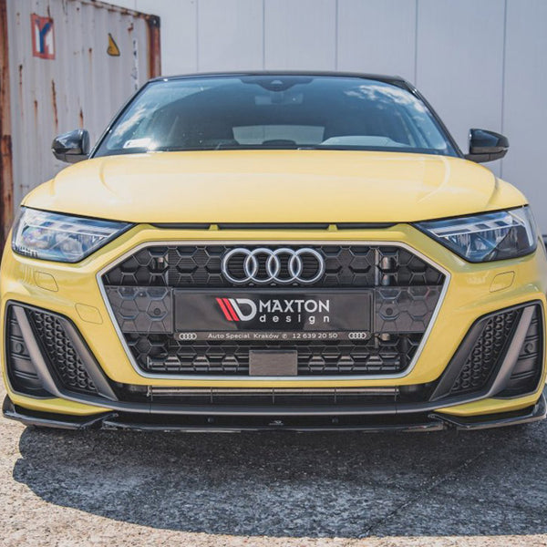A yellow Audi car is parked on pavement, featuring a prominent hexagonal grille with Maxton design branding. Shipping containers and a white building are visible in the background.