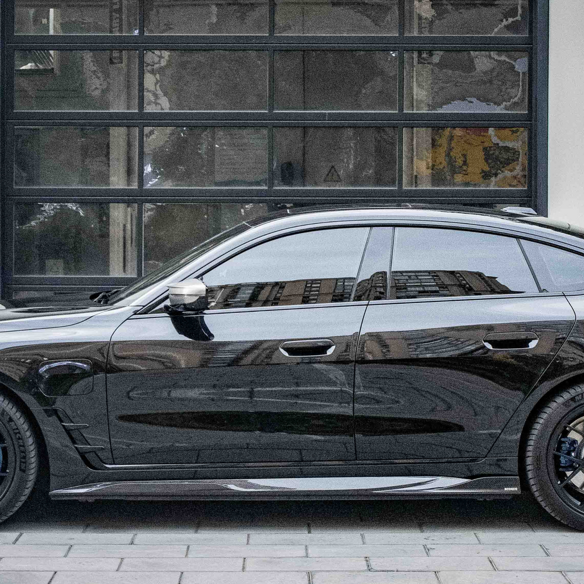 A sleek black car is parked on a cobblestone surface, reflecting nearby buildings, in front of a modern building with large, grid-like windows.