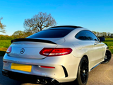 A white sports car is parked on a paved surface, featuring tinted windows and black wheels, with a clear blue sky and green fields in the background.