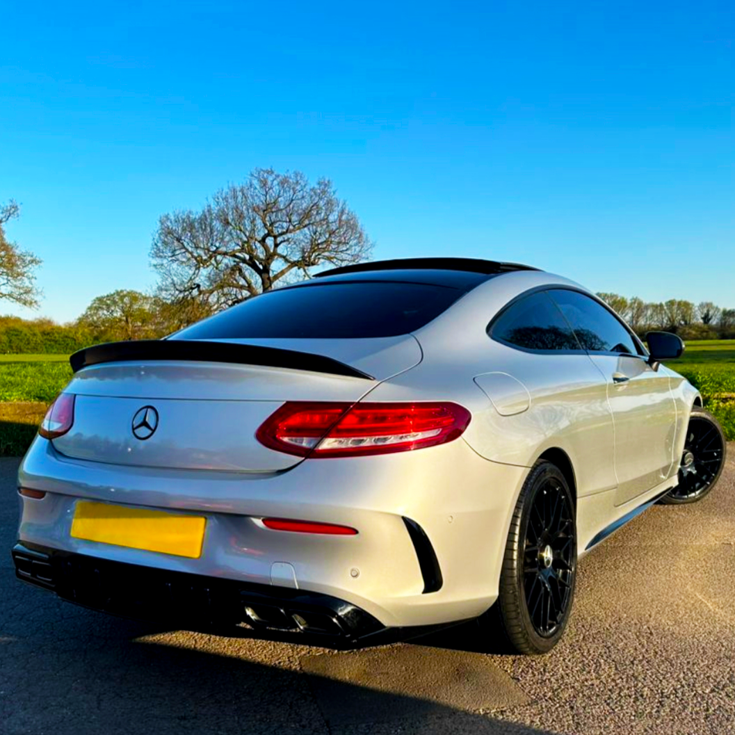 A white sports car is parked on a paved surface, featuring tinted windows and black wheels, with a clear blue sky and green fields in the background.