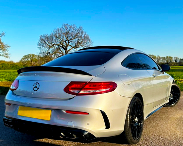 A white sports car is parked on a paved surface, featuring tinted windows and black wheels, with a clear blue sky and green fields in the background.