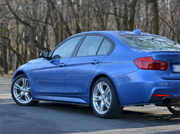 A blue sedan is parked on a paved surface, surrounded by leafless trees in a forested area.