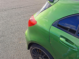 A green car with a rear spoiler is parked. It's situated in an open lot with other vehicles visible in the background under a clear sky.
