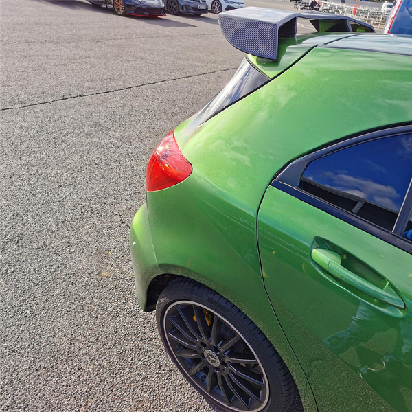 A green car with a rear spoiler is parked. It's situated in an open lot with other vehicles visible in the background under a clear sky.