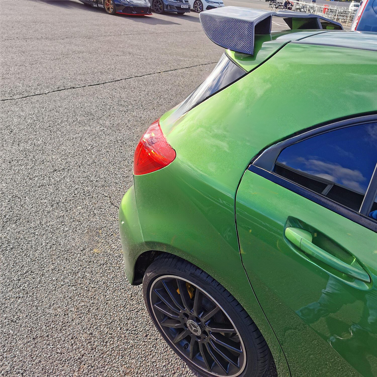 A green car with a rear spoiler is parked. It's situated in an open lot with other vehicles visible in the background under a clear sky.