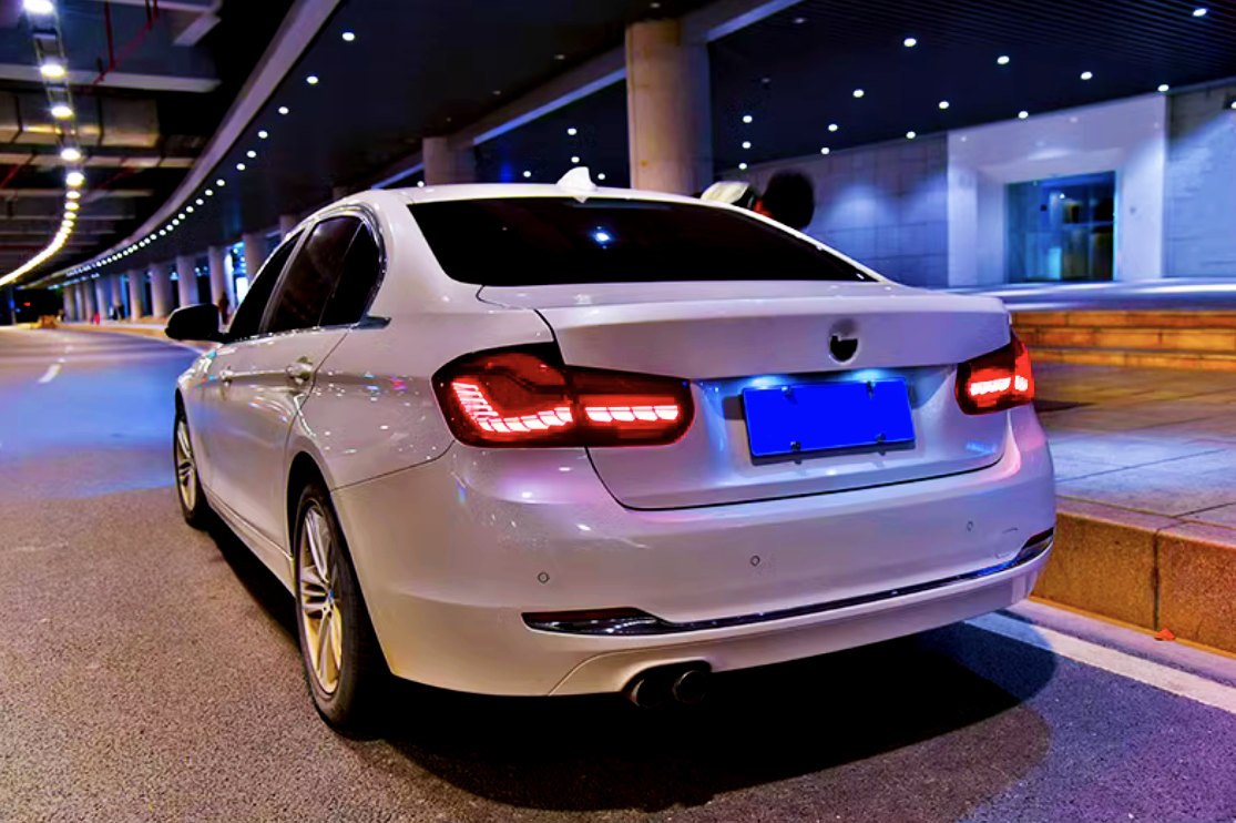 A white sedan is parked on a dimly lit street under a modern, roofed roadway. The taillights glow red, and nearby is a modern building entrance.