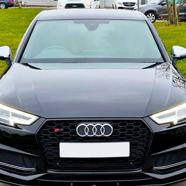 A black Audi car is parked on a road, facing forward. It features distinctive headlights and a front grille with the Audi logo. In the background, grassy areas and other parked cars are visible.