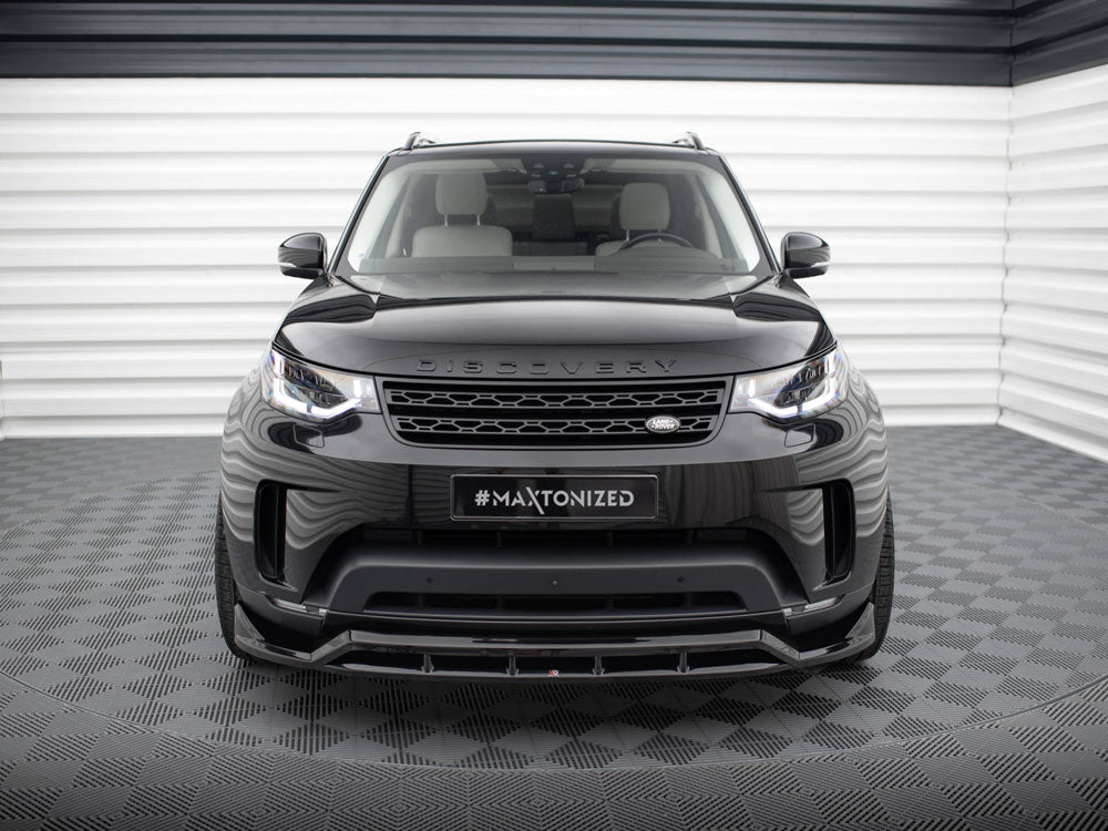 A black SUV, front view, with "DISCOVERY" on the hood and "#MAXTONIZED" on the license plate, parked on a checkered floor in a bright, striped indoor setting.