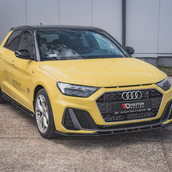 A yellow Audi car is parked on a concrete surface. The front grille features the Audi logo. Text on the grille reads "MAXTON design." A gray industrial building is in the background.