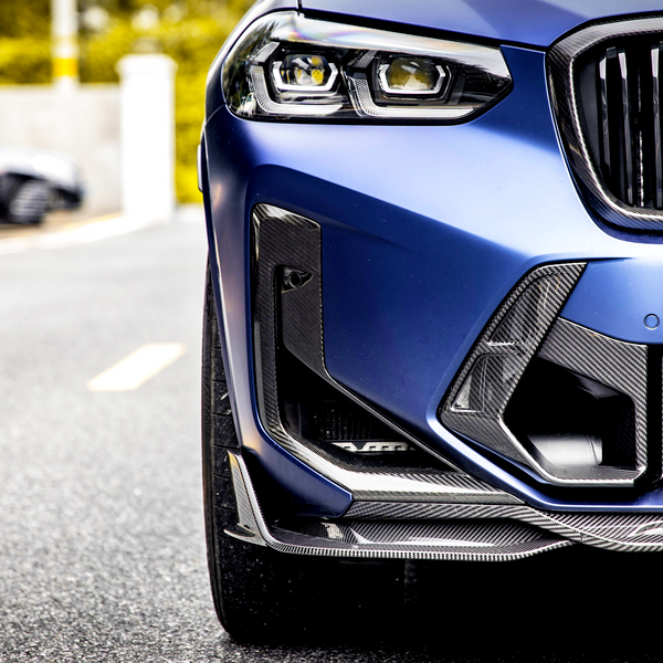 A close-up of a blue car's front detailing its grill and headlights, parked on a road lined with yellow markings, with blurred cars visible in the background.