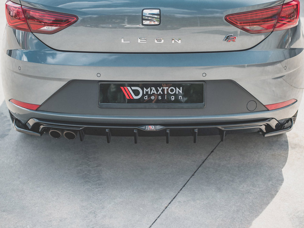 A gray car is parked with its rear bumper visible, featuring Maxton Design branding and dual exhausts. The setting is an outdoor concrete area.