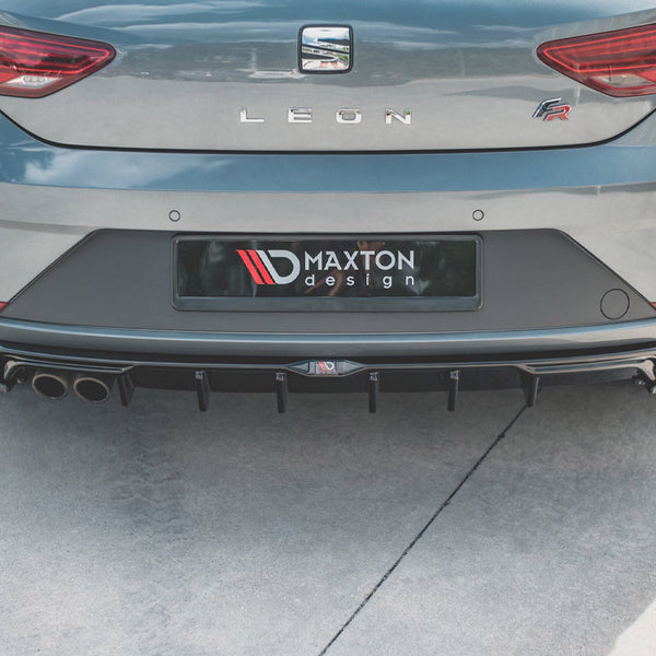 A gray car is parked with its rear bumper visible, featuring Maxton Design branding and dual exhausts. The setting is an outdoor concrete area.