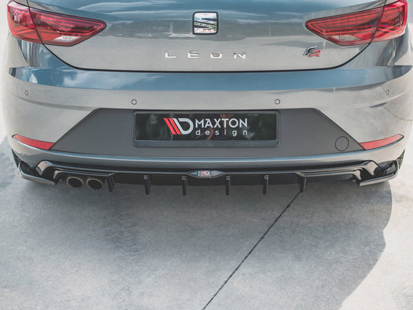 A gray car is parked with its rear bumper visible, featuring Maxton Design branding and dual exhausts. The setting is an outdoor concrete area.